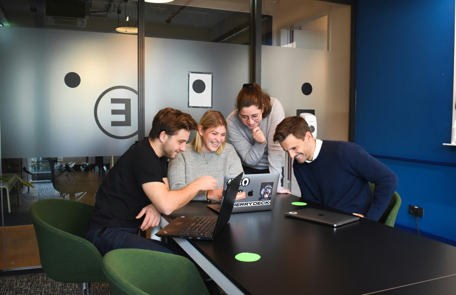 Vier collega’s werken samen rond laptops aan een tafel in een moderne kantoorruimte.
